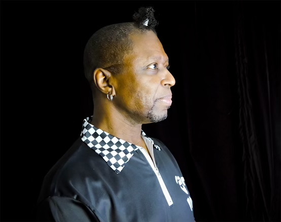 DJ Foxxy, a black man, is seen in profile fron the shoulders up. His hair is cropped very short except for a perfect little ponytail at the top of his forehead. He's wearing a black shirt with white piping an a checkerboard collar.